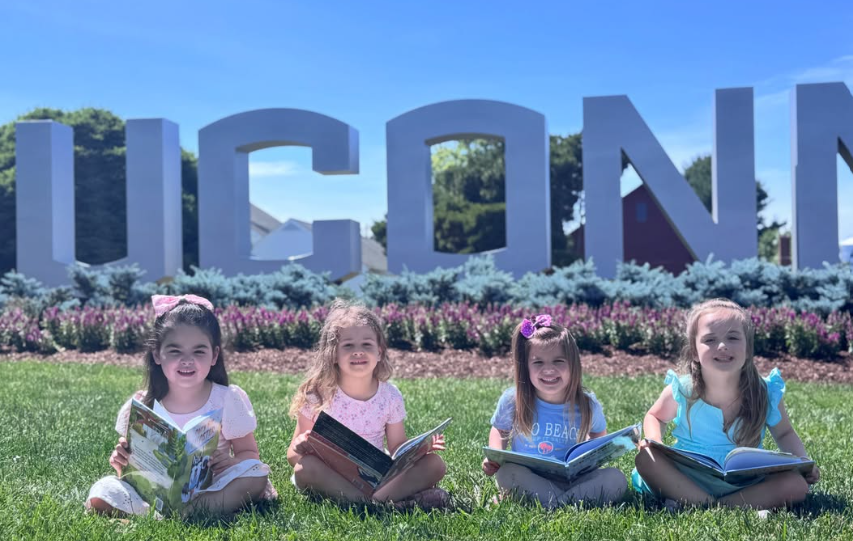 Four children reading books together on the grass in front of the UConn sign